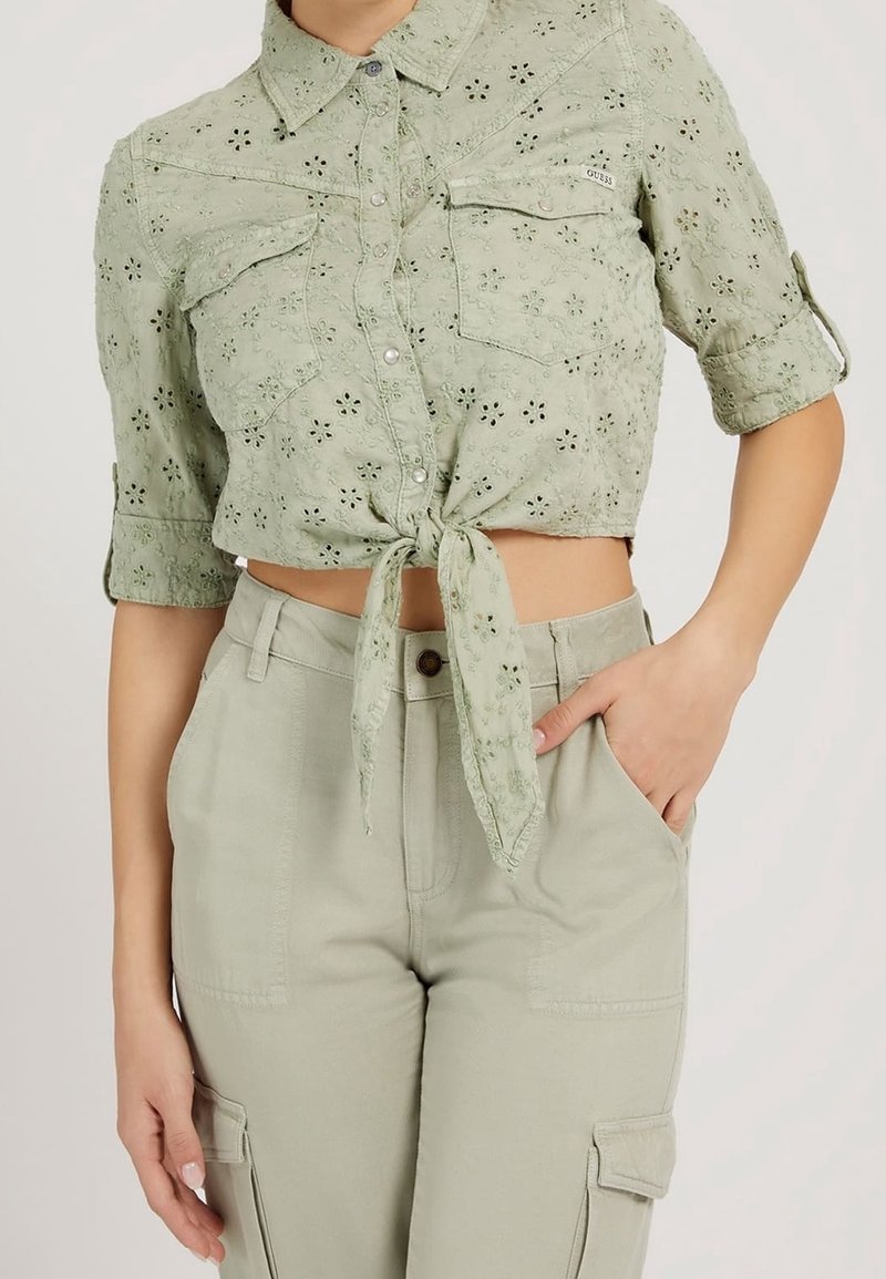 Woman wearing light green eyelet lace crop shirt tied at waist with rolled sleeves and light green high-waist cargo pants with pockets.