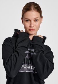 Jonge vrouw met lichtbruin haar draagt een zwarte hoodie, houdt de kraag met beide handen vast en kijkt recht in de camera.