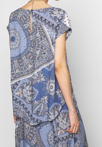 Sininen, kuvioitu mekko, jossa on paisley-tyylinen kuvio, lyhyet hihat, takana avainreikäyksityiskohta ja laskeutuva siluetti. Pehmeä kankaan rakenne.
