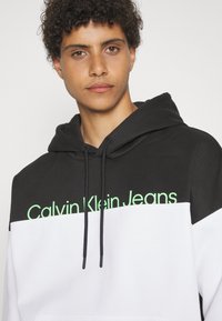 Čierno-biela mikina s kapucňou s zeleným logom "Calvin Klein Jeans". Obsahuje kapucňu na stiahnutie a kontrastné farebné bloky, vyrobená z mäkkého materiálu.