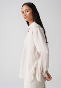 Lichtroze, gestreepte blouse met lange mouwen, een kraag en strikaccenten op de mouwen. De stof lijkt gestructureerd en heeft een relaxte pasvorm.