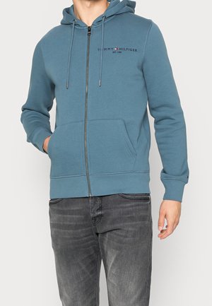 Mann trägt einen blauen Hoodie mit Reißverschluss und Fronttaschen sowie das „Tommy Hilfiger“-Logo, kombiniert mit ausgewaschenen schwarzen Jeans.