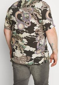 Mies, kiertänyt tumman kuviollisen paidan, jossa on lintuja, käärmeitä ja pilviä, harmaiden housujen kanssa, nähty selkäpuolelta valkoista taustaa vasten.