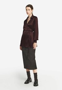 Burgundy satin omlottopp med lång ärm och knyt detaljer, kombinerad med en mörk kjol. Modellen har svarta skor och bruna strumpor.