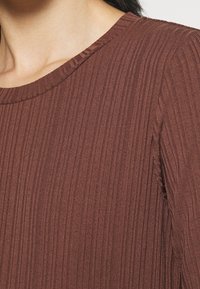 Brązowa bluzka z prążkowanej tkaniny, z okrągłym dekoltem i teksturowanym materiałem. Materiał wydaje się miękki, z pionowymi liniami dla dodatkowego wymiaru.
