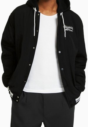 Mężczyzna w czarnej kurtce z kapturem z białymi sznureczkami, z napisem "Philadelphia" na piersi, białą koszulką pod spodem i czarnych spodniach z paskami.