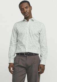 Camisa blanca de botón con un ligero patrón floral en azul y verde, combinada con pantalones grises y un cinturón negro trenzado.