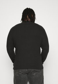 Chemise noire à manches longues avec une finition texturée, présentant un col rond et un ourlet côtelé. Le design est simple, sans logos ou motifs visibles.