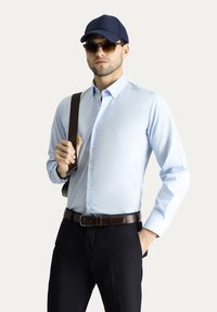 Camicia azzurra chiara con colletto, indossata con pantaloni scuri e una cintura marrone. Il modello tiene una borsa a spalla marrone e indossa un cappellino blu navy e occhiali da sole.