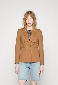 Blazer marron à double boutonnage avec des boutons dor�és, coupe ajustée et revers élégants, porté sur un haut en dentelle noire et associé à un short en denim.