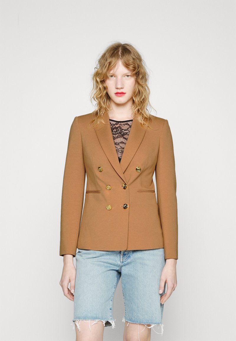Blazer marrón de doble botonadura con botones dorados, ajuste entallado y solapas elegantes, llevado sobre una blusa de encaje negra y combinado con pantalones cortos de mezclilla.