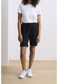 Camiseta de algodón blanca combinada con pantalones cortos negros a la rodilla con cinturilla elástica y bolsillos laterales, llevada con zapatillas blancas.