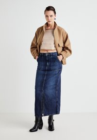 Chaqueta bomber corta marrón, top de punto a rayas y falda maxi de mezclilla oscura con una abertura frontal. Llevado con botas negras de tacón.