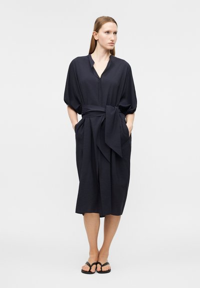 Γυναίκα με μαύρο midi φόρεμα με ζώνη δεμένη και κοντά φουσκωτά μανίκια, στέκεται με τα χέρια στις τσέπες, φοράει μαύρα σανδάλια σε απλό φόντο.