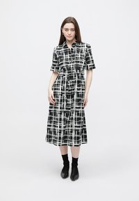 Vestido midi a cuadros en blanco y negro con mangas cortas, cierre frontal de botones, cintura con cordón ajustable y falda fluida. Lleva botas negras hasta el tobillo.