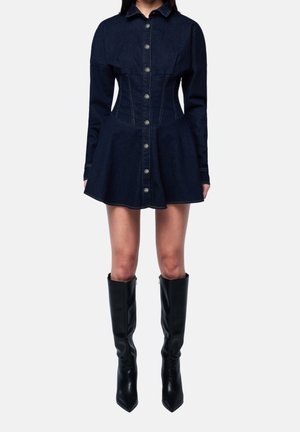 Kobieta w ciemnej dżinsowej sukience na guziki z długimi rękawami, rozkloszowaną spódnicą oraz czarnymi, zakolanówkami z ostrym noskiem na tle jednolitego tła.