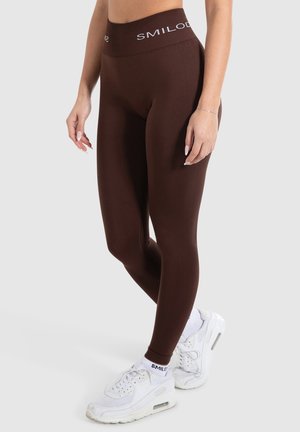 Brune leggings med høj talje og hvid "SMILOD" branding. Båret med hvide træningssko, der viser et glat, strækbart materiale.