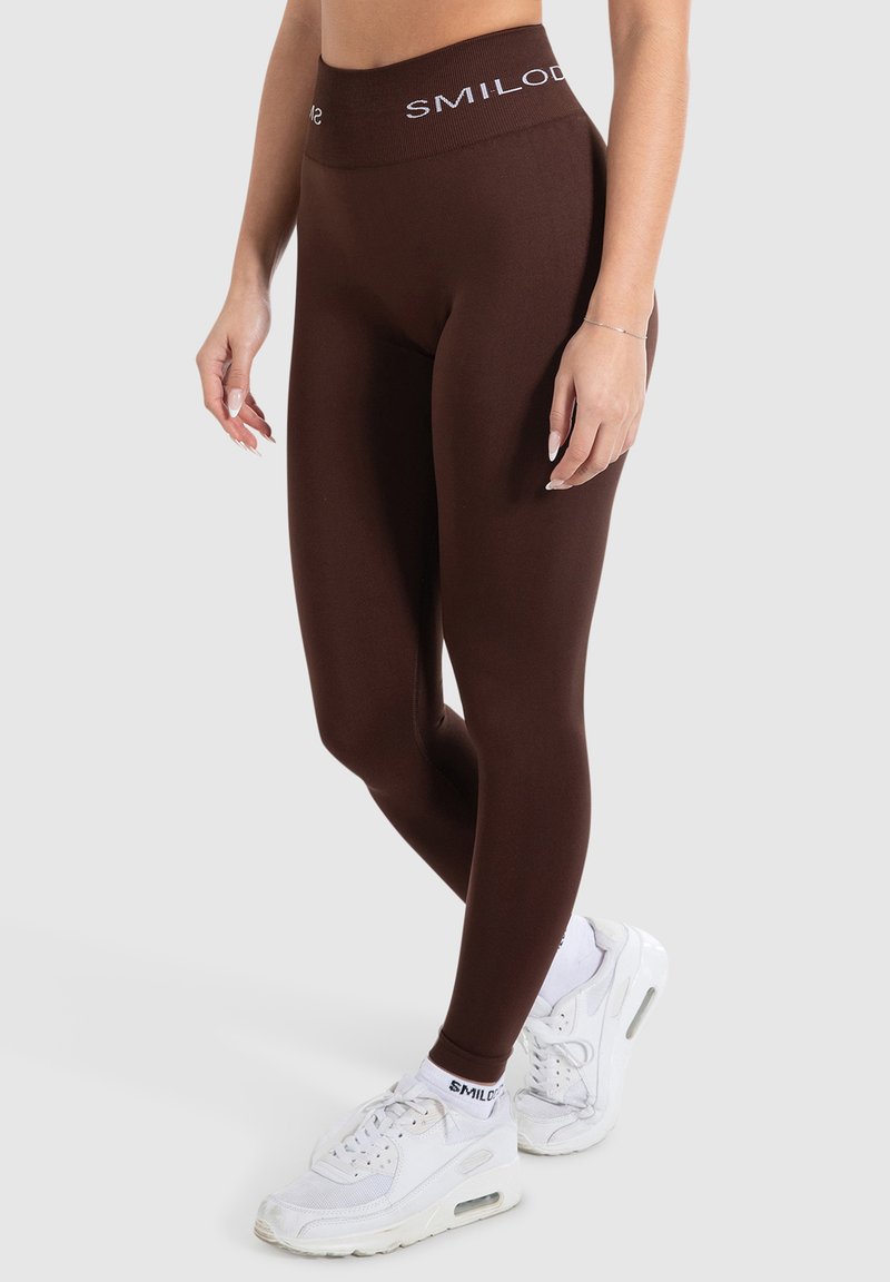 Leggings marroni con una vita alta caratterizzati dal marchio bianco "SMILOD". Indossati con scarpe da ginnastica bianche, che mettono in mostra un tessuto liscio e elasticizzato.
