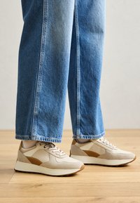 Beige Sneaker mit Wildleder- und Netzpaneelen, akzentuiert durch hellbraune Details, runde Zehen und weiße Sohle, kombiniert mit blauen Jeans.