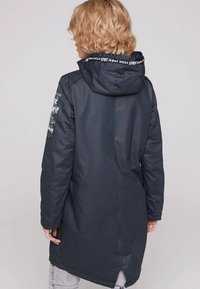 Blondyn w długim, ciemnym płaszczu z kapturem, z białym napisem na krawędzi kaptura i lewym rękawie, stojący na jednolitym jasnym tle.