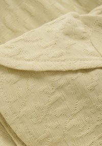 Knittrikå i ljusgul med ett texturerat mönster av sammanlänkade former. Mjuk, slät yta, vikt för att visa djup och detalj.