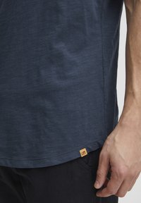 Tricou bleu marin cu mânecă scurtă, textură moale, cu margine rotunjită, având un mic etichetă portocalie pe partea de jos. Purtați cu pantaloni de culoare închisă.