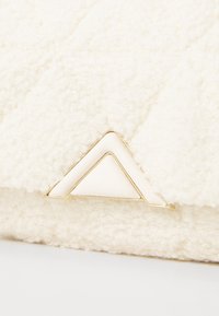 Hvid struktureret taske med en fuzzy overflade, et gyldent trekantet emblem og en slank hvid detalje nær lukningen.