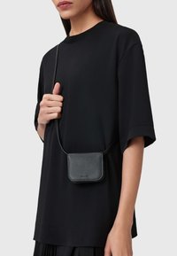 Borsetta a tracolla in pelle nera con una texture liscia e design minimalista, indossata sopra una camicia nera larga. Dettaglio di chiusura semplice visibile.