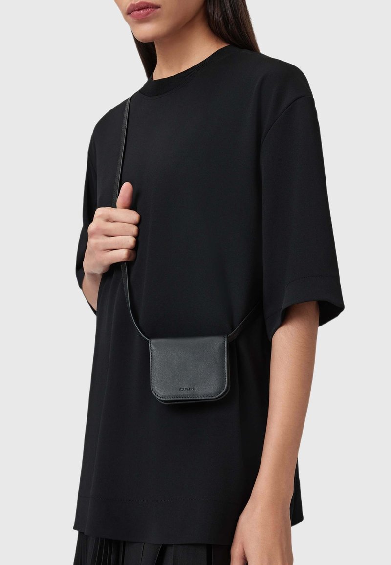 Borsetta a tracolla in pelle nera con una texture liscia e design minimalista, indossata sopra una camicia nera larga. Dettaglio di chiusura semplice visibile.