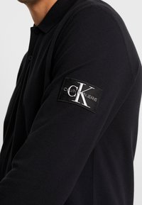 Muž v černém polo tričku s dlouhým rukávem s nášivkou loga Calvin Klein Jeans na horní části rukávu na jednobodovém pozadí.