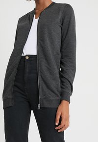 Donna che indossa una giacca cardigan zip-up grigio scuro sopra una maglietta bianca a V e jeans neri a vita alta, in piedi con la mano destra rilassata accanto.