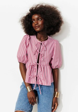 Femme aux cheveux bouclés volumineux portant un chemisier rose rayé avec des liens à l'avant, un jean bleu, des bracelets en or et un collier chaîne, debout devant un fond blanc.