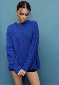 Modro pleten pulover z okroglim izrezom, ohlapne oblike in rebrastimi manšetami; nošen z temnimi kratkimi hlačami na enobarvni svetlo modri podlagi.