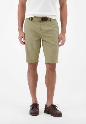 Mann trägt beige Shorts mit einem braunen Gürtel und dunkelbraunen Schnürschuhen, steht vor einem schlichten weißen Hintergrund.
