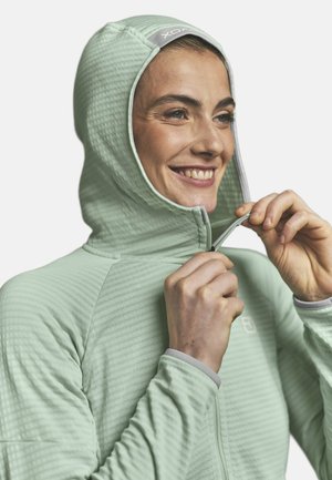 Woman smiling while zipping up a light green textured hooded jacket, shown from the chest up on a plain background.