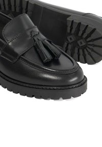 Black leather loafers with a textured surface, featuring tassels on the front and a rugged black sole with pronounced treads.