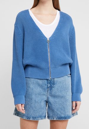 Kvinde iført en blå strikket cardigan med lynlås over en hvid top og lyseblå denimshorts, stående mod en ensfarvet baggrund.