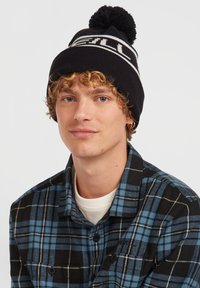 Black knit beanie with a white and light blue logo and a pom-pom on top, worn by a person in a blue plaid shirt with a black background.