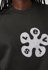 Gros plan sur un sweat-shirt foncé avec une forme de fleur blanche abstraite et les lettres « l », « e », « v », « i », « s » sur la poitrine.