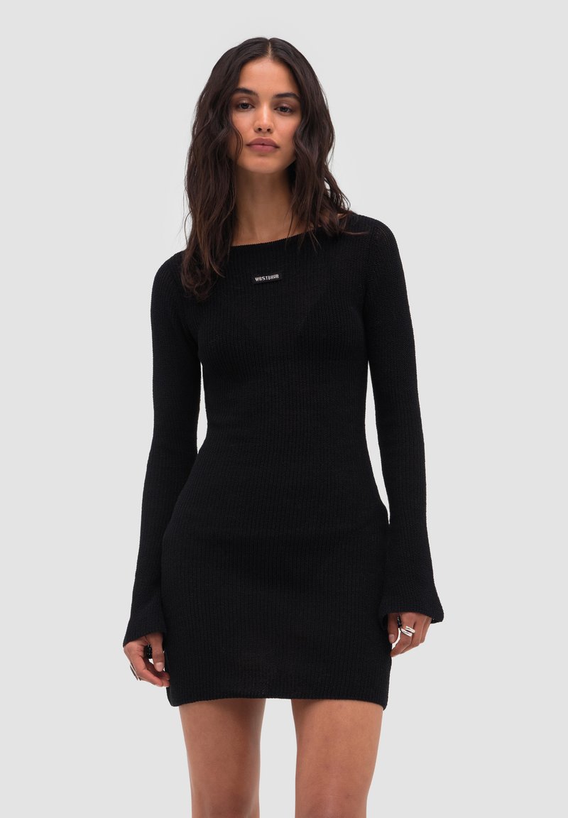 Femme aux longs cheveux bruns portant une robe mini noire ajustée en maille à manches longues, sur un fond gris clair.