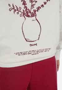 Biele mikina s bordovým obrysom vázy s kvetinovými akcentmi, doplnená o popisný text na spodnej časti. Nosí sa s bordovými nohavicami.