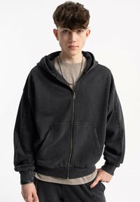 Czarna bluza z kapturem, z suwakiem, kieszenią kangurką i ściągaczami przy mankietach. Nosi się na warstwie beżowej koszuli. Miękka, teksturowana tkanina.