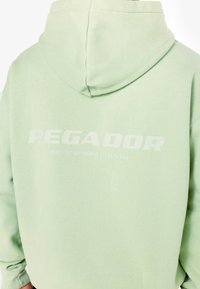 Svijetlozelena dukserica s kapuljačom koja ima mekanu teksturu. Na leđima se nalazi naziv branda "PEGADOR" i fraza "CERTIFICIRANI AUTENTIČNI ULIČNI MODA."