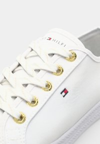 White sneaker with white laces, gold eyelets, and small red, white, and blue logo on the side and "Tommy Hilfiger" text on the tongue.