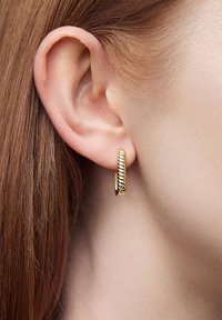 Złoty kolczyk w kształcie obręczy z kręconym wzorem, gładką krawędzią i błyszczącym wykończeniem, zaprezentowany w zbliżeniu na ucho.