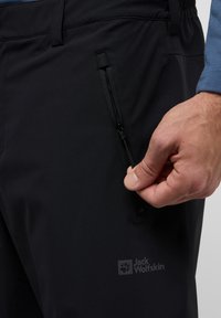 Zwarte outdoorbroek met een ritszak op de bovenbeen en een logo. Het materiaal is lichtgewicht en heeft een gladde textuur.
