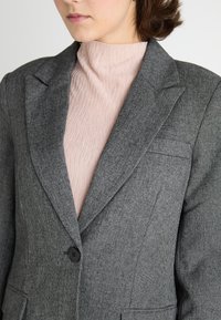 Grijze blazer van textuurstof met notch revers, een enkele knoopsluiting en een borstzak, gedragen over een lichtroze top.