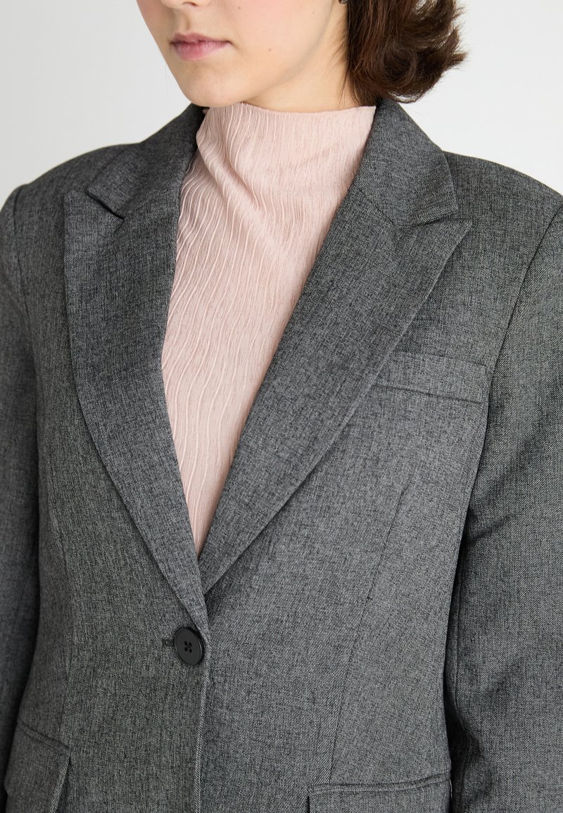 Blazer grigio in tessuto texturizzato con revers a scollatura, chiusura con un solo bottone e taschino sul petto, indossato sopra una maglietta rosa chiaro.