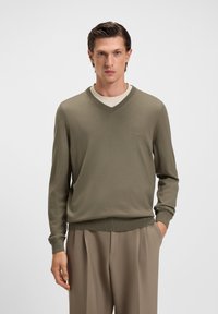 Mann in olivgrünem V-Ausschnitt-Pullover über beigem Hemd mit beigen Hosen, steht mit einer Hand in der Tasche vor einem einfarbigen Hintergrund.