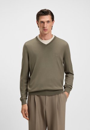 Mann in olivgrünem V-Ausschnitt-Pullover über beigem Hemd mit beigen Hosen, steht mit einer Hand in der Tasche vor einem einfarbigen Hintergrund.